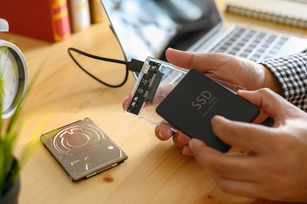 Osoba drži SSD disk u eksternom kućištu pored HDD diska na drvenom stolu, sa laptopom u pozadini
